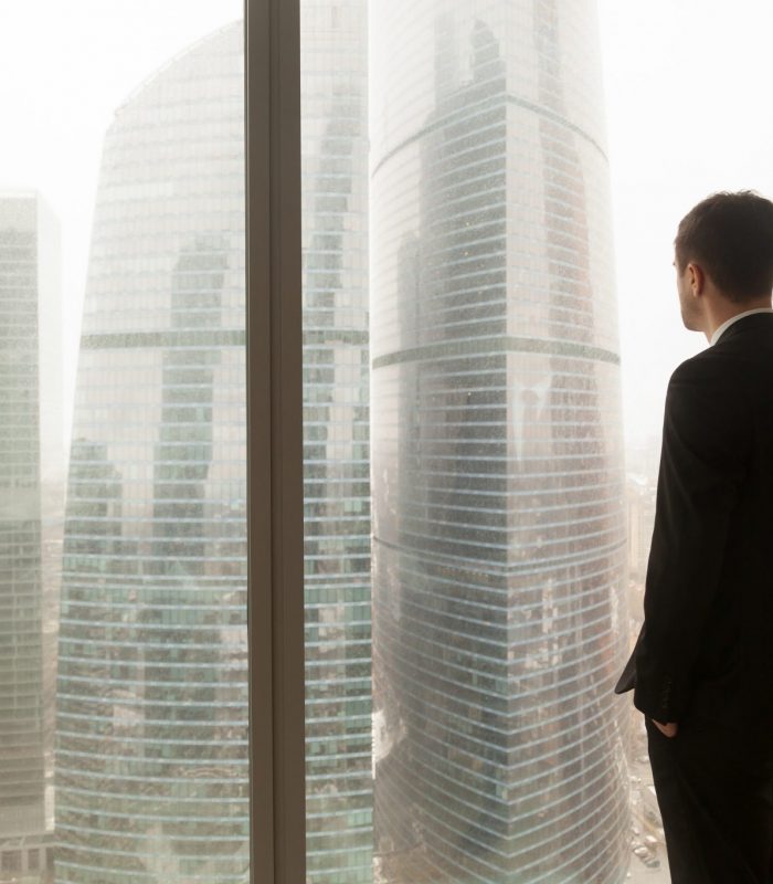 Man in business suit standing with hands in pants pockets looking through window on city landscape with skyscrapers. Businessman thinking about perspectives, dreaming of company success. Back view